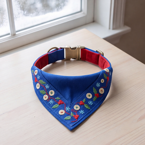 A blue pet bandana collar with red lining and embroidered floral details, displayed on a light wood table near a window.
