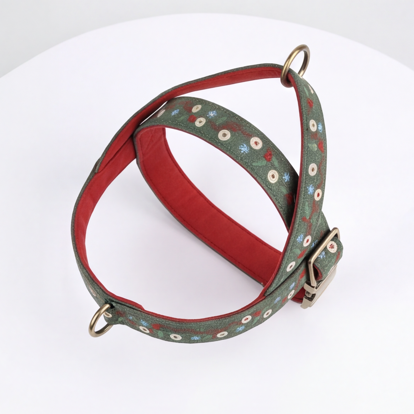 A green patterned dog harness with a red interior lining, brass hardware, and a buckle, displayed on a white surface.