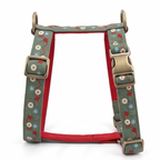 A dog harness with a sage green canvas exterior featuring a whimsical pattern of red hearts, blue snowflakes, and white buttons, accented by a bright red interior lining and antique brass hardware.