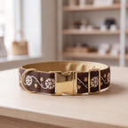 A brown patterned dog collar with cream floral and vine motifs and a shiny gold buckle, displayed on a light wooden surface with a softly blurred indoor background.