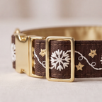 A close-up of a brown woven fabric collar with white embroidered snowflakes and gold floral details, featuring a gold-tone metal buckle.