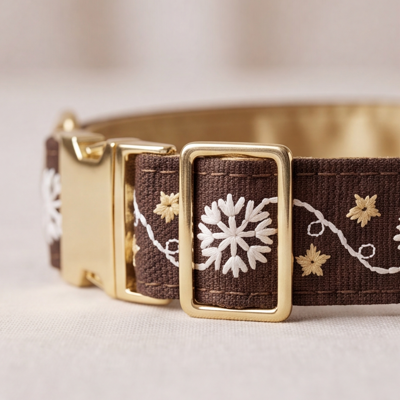 A close-up of a brown woven fabric collar with white embroidered snowflakes and gold floral details, featuring a gold-tone metal buckle.