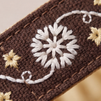 Close-up of a brown crocheted or knitted textile with white embroidered floral motifs and small beige flowers, shown diagonally against a softly blurred warm background.