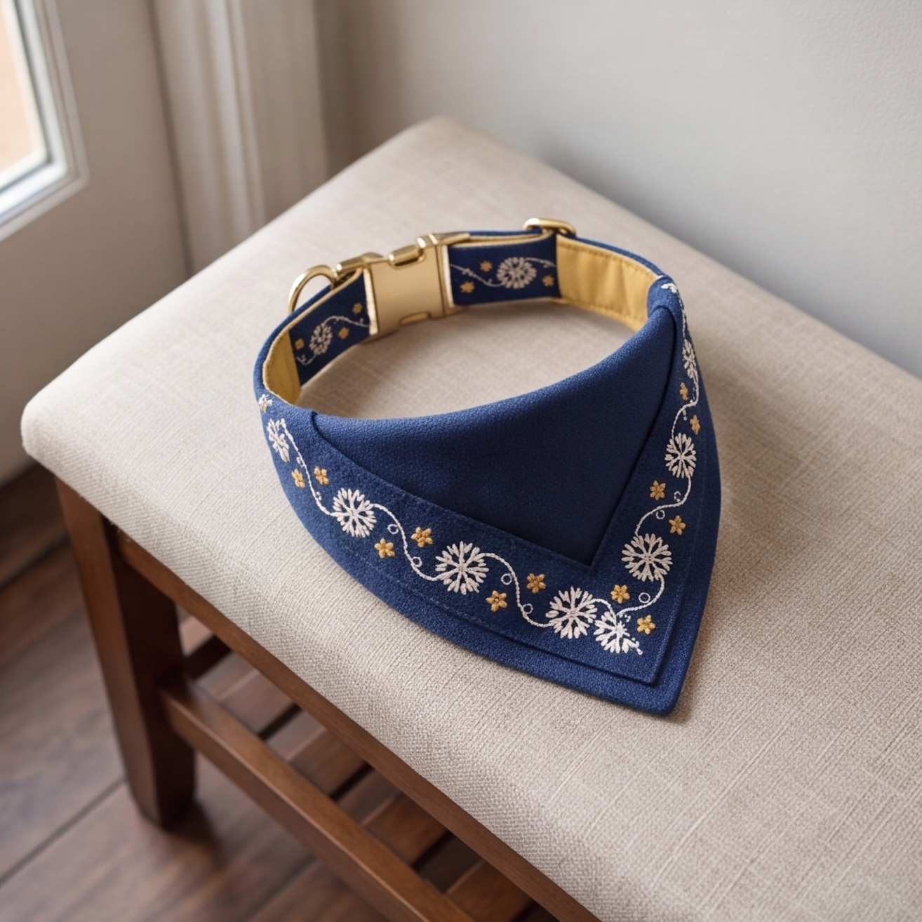A navy blue pet bandana with white floral embroidery and gold accents, displayed on a beige upholstered wooden bench.