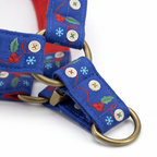 A close-up of a blue dog harness with embroidered holly and berry patterns, brass hardware, and red inner lining, shown on a white background.