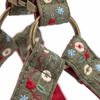 Close-up of a woven fabric pet harness strap with embroidered floral details in red, blue, and green on an olive green base, featuring brass-colored metal rings and a metal clasp.