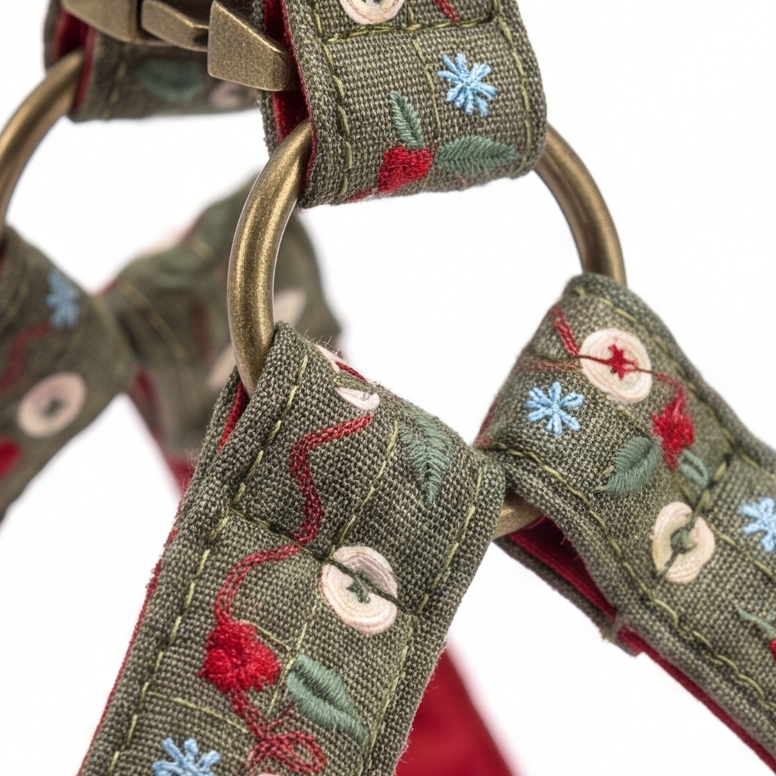 Close-up of a woven fabric pet harness strap with embroidered floral details in red, blue, and green on an olive green base, featuring brass-colored metal rings and a metal clasp.