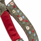 Close-up of a woven fabric strap, likely a dog collar, featuring olive green outer fabric with embroidered red and blue flowers, cream buttons, and green leaves, over a red satin inner lining.