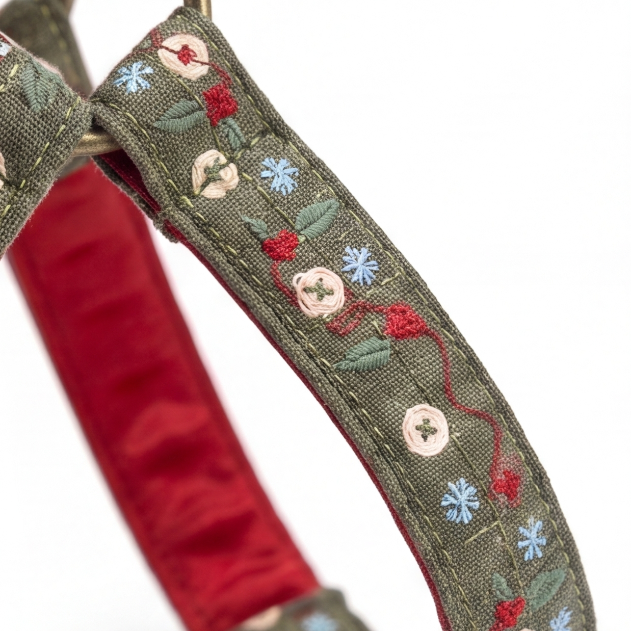 Close-up of a woven fabric strap, likely a dog collar, featuring olive green outer fabric with embroidered red and blue flowers, cream buttons, and green leaves, over a red satin inner lining.