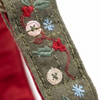 Close-up of an olive green embroidered fabric strap or trim featuring red floral motifs, green leaves, light blue snowflake-like stitches, and two round beige buttons with green centers, set against a white background with a red fabric edge visible on the left.
