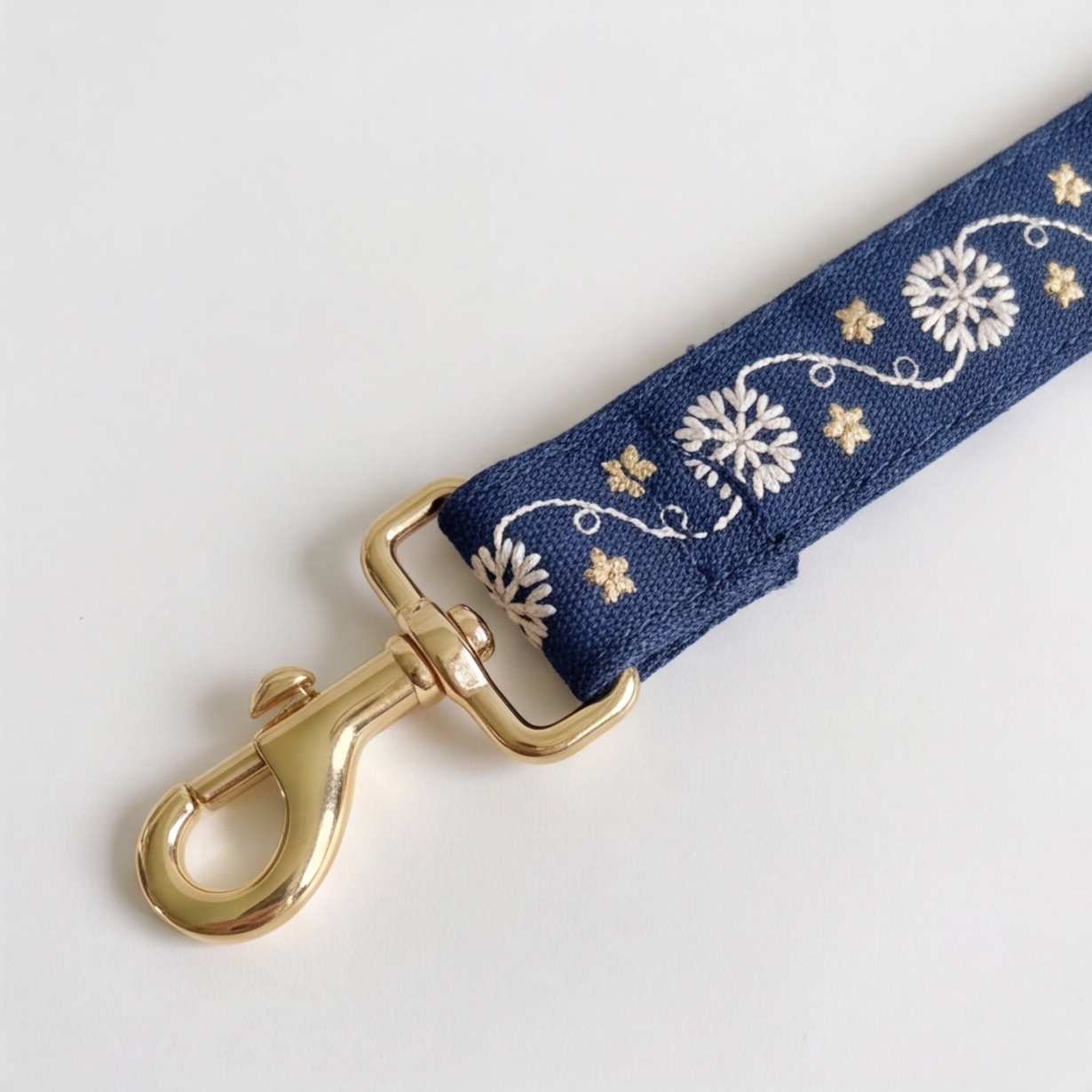 A close-up of a navy blue fabric strap with white floral embroidery and gold-tone metal hardware, including a lobster clasp, on a plain light background.
