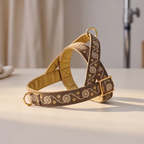 A brown and gold embroidered dog harness with floral patterns and gold hardware, displayed on a light beige surface with a softly blurred background.
