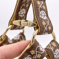 Close-up of a brown fabric strap with white floral embroidery and gold-tone hardware, held by a hand against a light background.