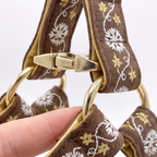 Close-up of a brown fabric strap with white floral embroidery and gold-tone hardware, held by a hand against a light background.
