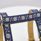 A close-up of a navy blue pet harness with white embroidered floral and star patterns, featuring gold-toned metal rings and a tan interior lining.