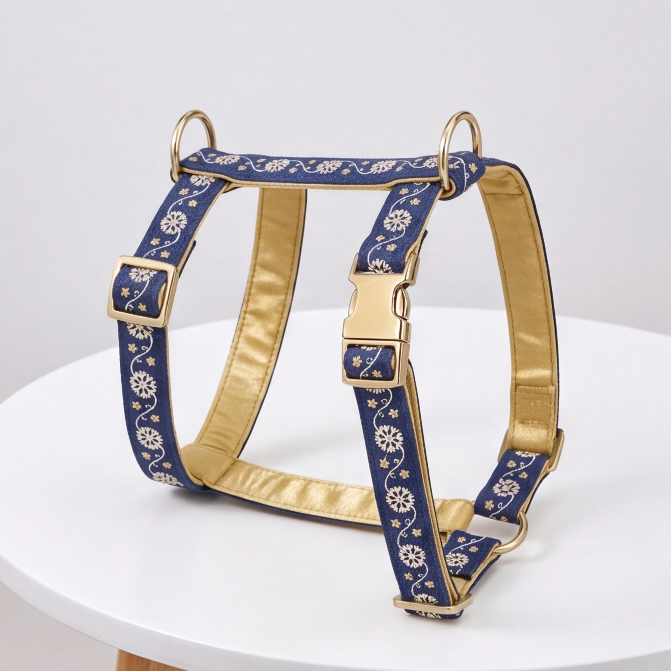 A blue and gold dog harness with a floral pattern, displayed on a white round table against a light gray background.