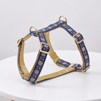 A navy blue dog harness with gold floral embroidery and gold-toned metal hardware, displayed on a white round table against a light gray background.