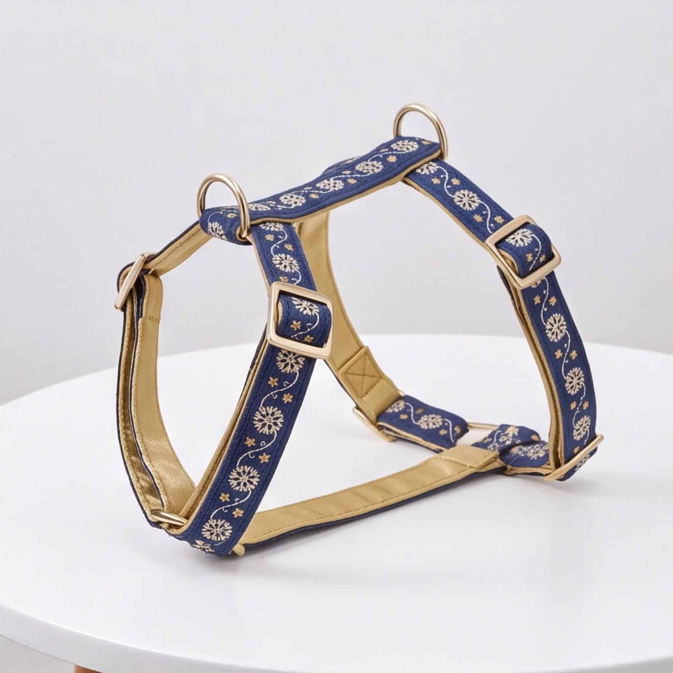 A navy blue dog harness with gold floral embroidery and gold-toned metal hardware, displayed on a white round table against a light gray background.