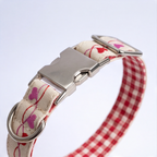 A close-up of a dog collar with a red and white gingham pattern, a cream-colored woven fabric side printed with pink and red hearts, and a silver-tone metal buckle and D-ring.