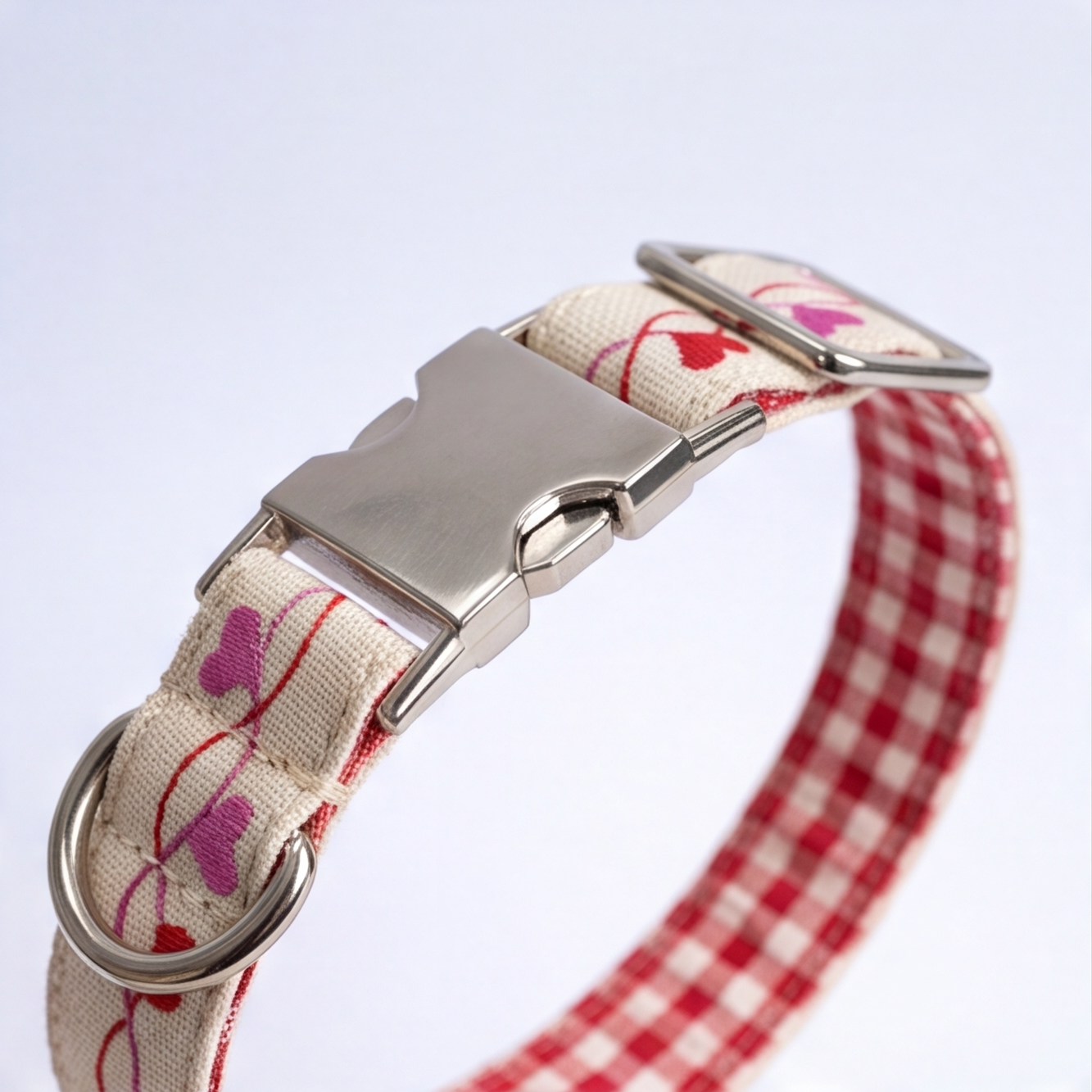 A close-up of a dog collar with a red and white gingham pattern, a cream-colored woven fabric side printed with pink and red hearts, and a silver-tone metal buckle and D-ring.