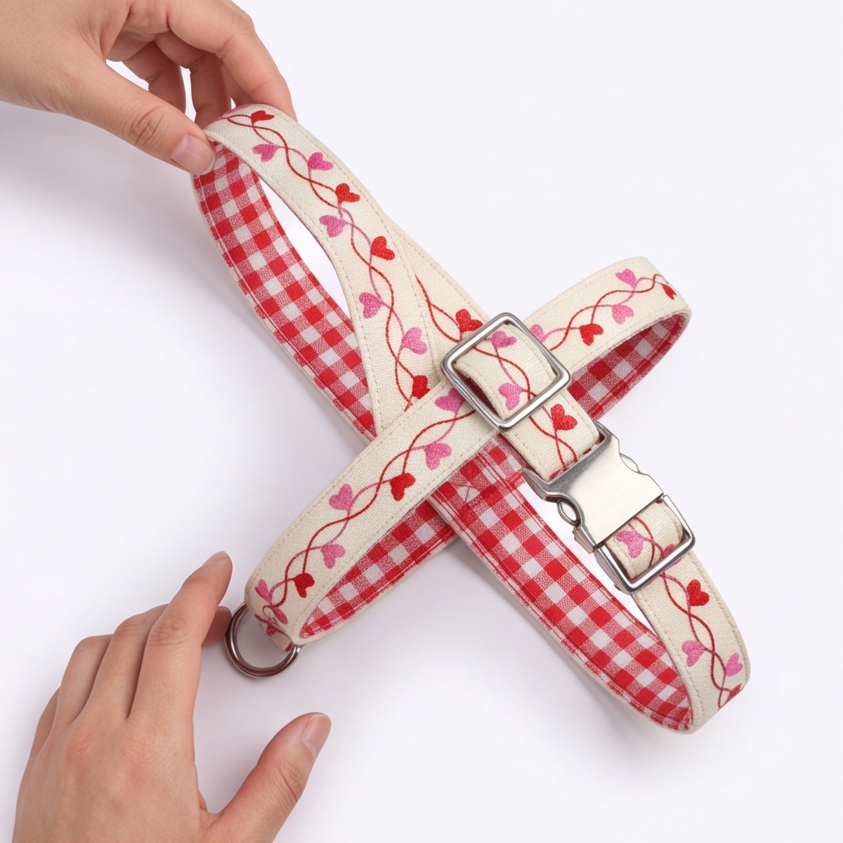 A dog harness with red and white gingham check fabric, decorated with red and pink heart-shaped vines, held by two hands against a plain white background.