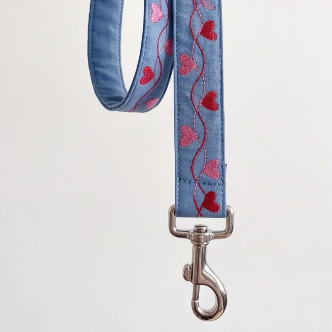 A light blue denim-style strap with embroidered red and pink hearts and wavy lines, finished with a silver metal swivel clasp, shown against a plain light background.