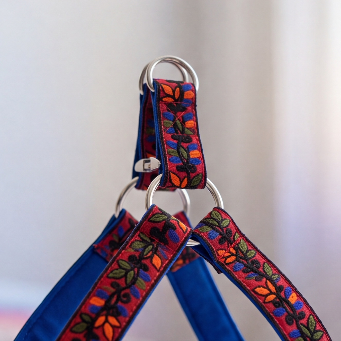 A close-up of a pet harness with a vibrant multicolor floral embroidered strap and solid blue fabric straps, attached to silver metal rings.