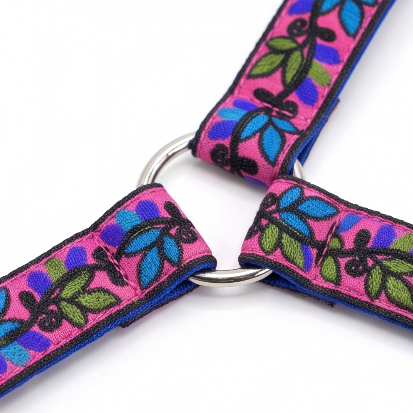 A close-up of a pink pet harness strap with a colorful embroidered floral pattern in blue, green, and purple, featuring a silver metal D-ring connector.