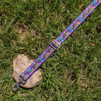 A colorful dog leash with a repeating floral pattern lies on green grass next to a small stone. The leash has a blue-purple base with pink, green, and orange flowers, silver metal hardware, and a matching buckle.