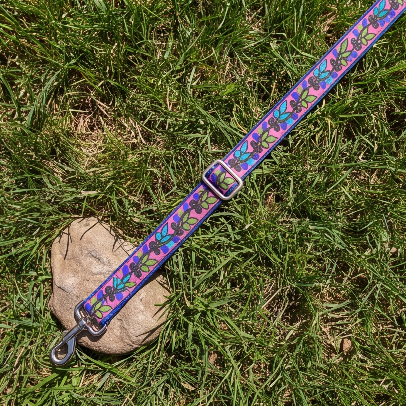 A colorful dog leash with a repeating floral pattern lies on green grass next to a small stone. The leash has a blue-purple base with pink, green, and orange flowers, silver metal hardware, and a matching buckle.