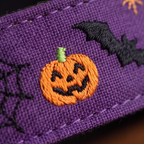 Close-up of a purple embroidered fabric featuring a jack-o'-lantern pumpkin, black bat, spiderweb, and orange spider detail.