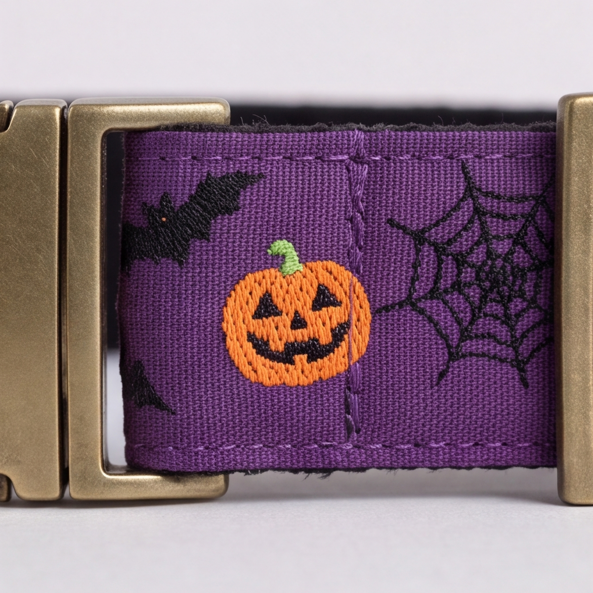 A close-up of a purple woven strap with a gold-tone buckle, featuring embroidered Halloween motifs including a jack-o'-lantern, black bats, and a spiderweb.