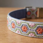 A close-up of a woven pet collar with a navy blue interior and a detailed embroidered exterior. The exterior features a geometric pattern in light blue, green, and white, accented with pink and orange embroidered flowers.