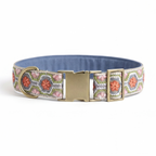 A patterned fabric dog collar with a blue interior, featuring a pink, green, and white geometric design with orange floral motifs, and a gold-tone metal buckle and D-ring, shown on a white background.