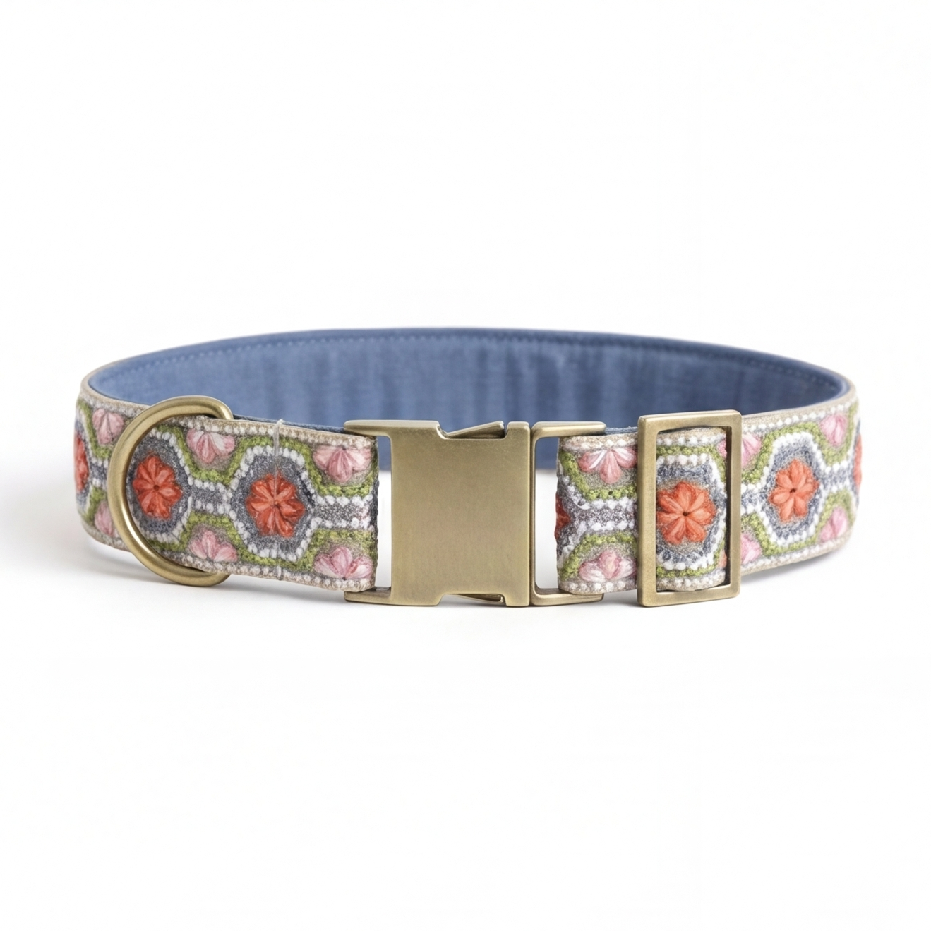 A patterned fabric dog collar with a blue interior, featuring a pink, green, and white geometric design with orange floral motifs, and a gold-tone metal buckle and D-ring, shown on a white background.