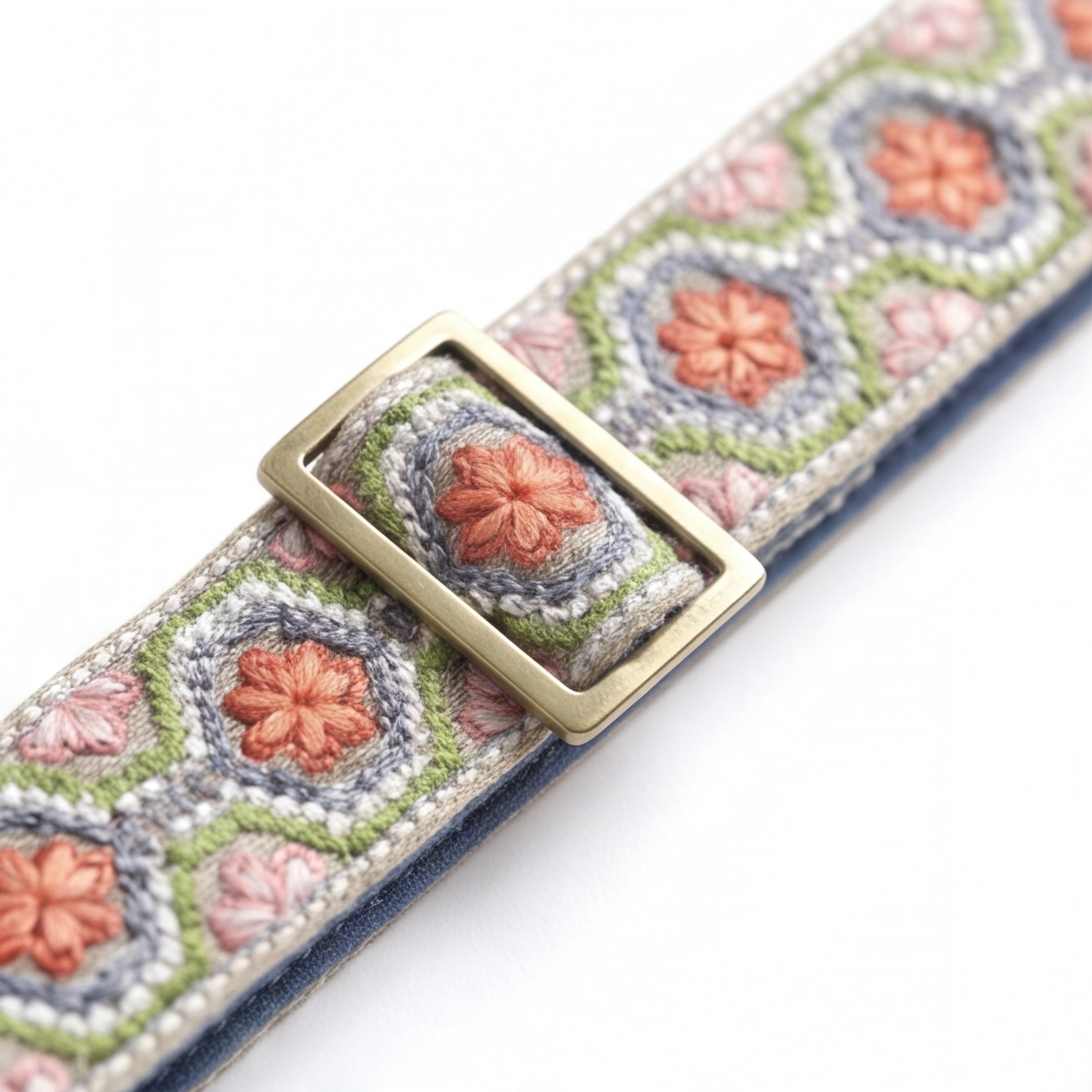 A close-up of a woven fabric belt with a geometric pattern featuring pink and orange flowers, green and blue-green outlines, and a brass buckle.