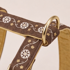 Close-up of a brown fabric harness with white floral embroidery, gold star motifs, and a gold metal D-ring on a light beige background.