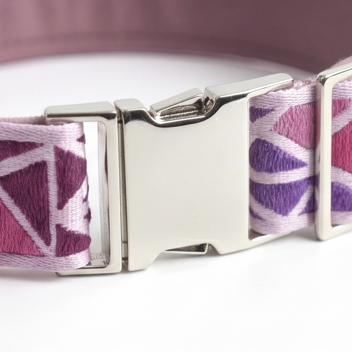 A close-up of a woven pet collar with a geometric pattern in purple, magenta, and white, featuring a silver metal buckle.