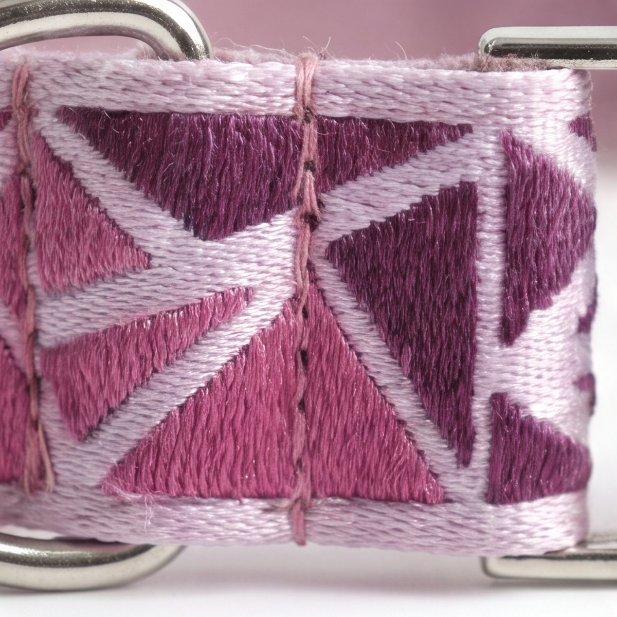 Close-up of a woven or embroidered strap with a geometric triangle pattern in pink, mauve, and pale lavender, with silver metal hardware visible at the corners.