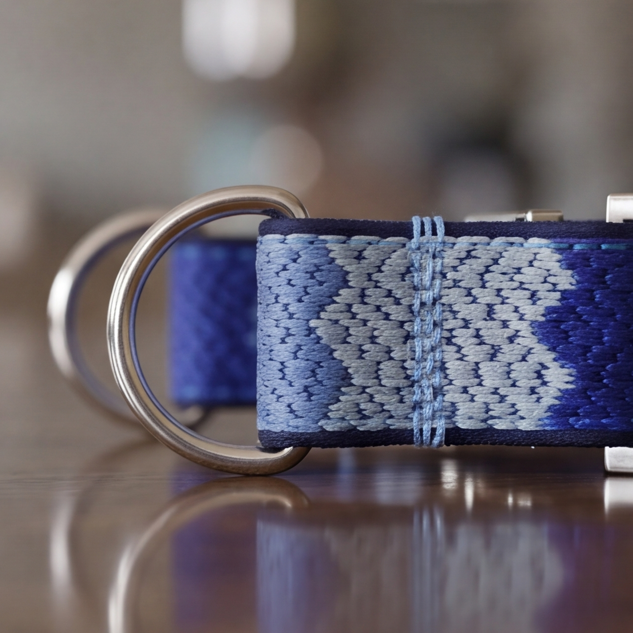 A close-up of a woven dog collar with a blue and white geometric pattern, featuring silver metal D-rings, resting on a reflective wooden surface.
