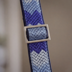 A close-up of a woven strap with a geometric chevron pattern in shades of blue, purple, and white, featuring a silver-toned metal buckle.