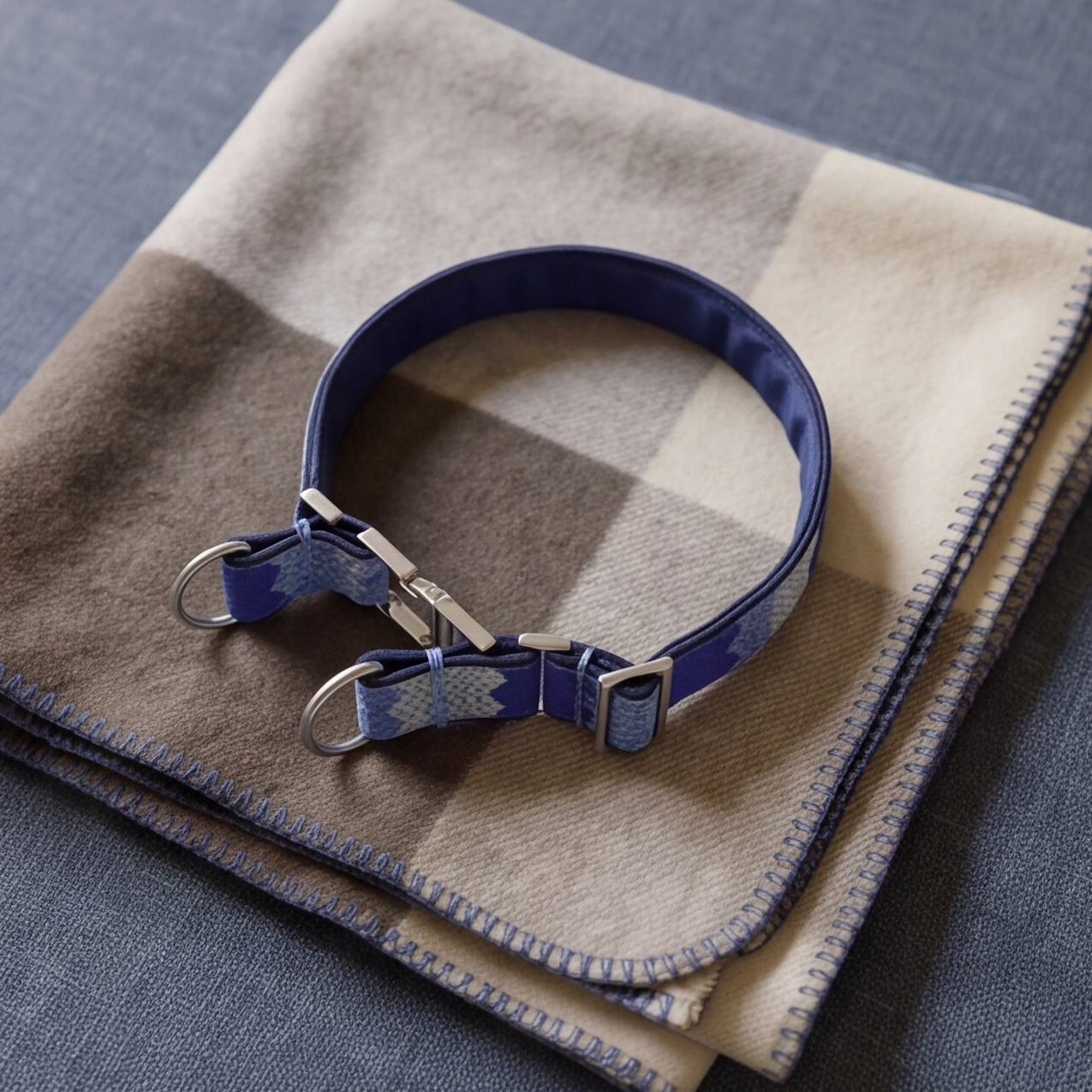 A blue patterned dog collar with silver hardware, displayed on a folded beige and brown plaid blanket over a blue-gray fabric background.