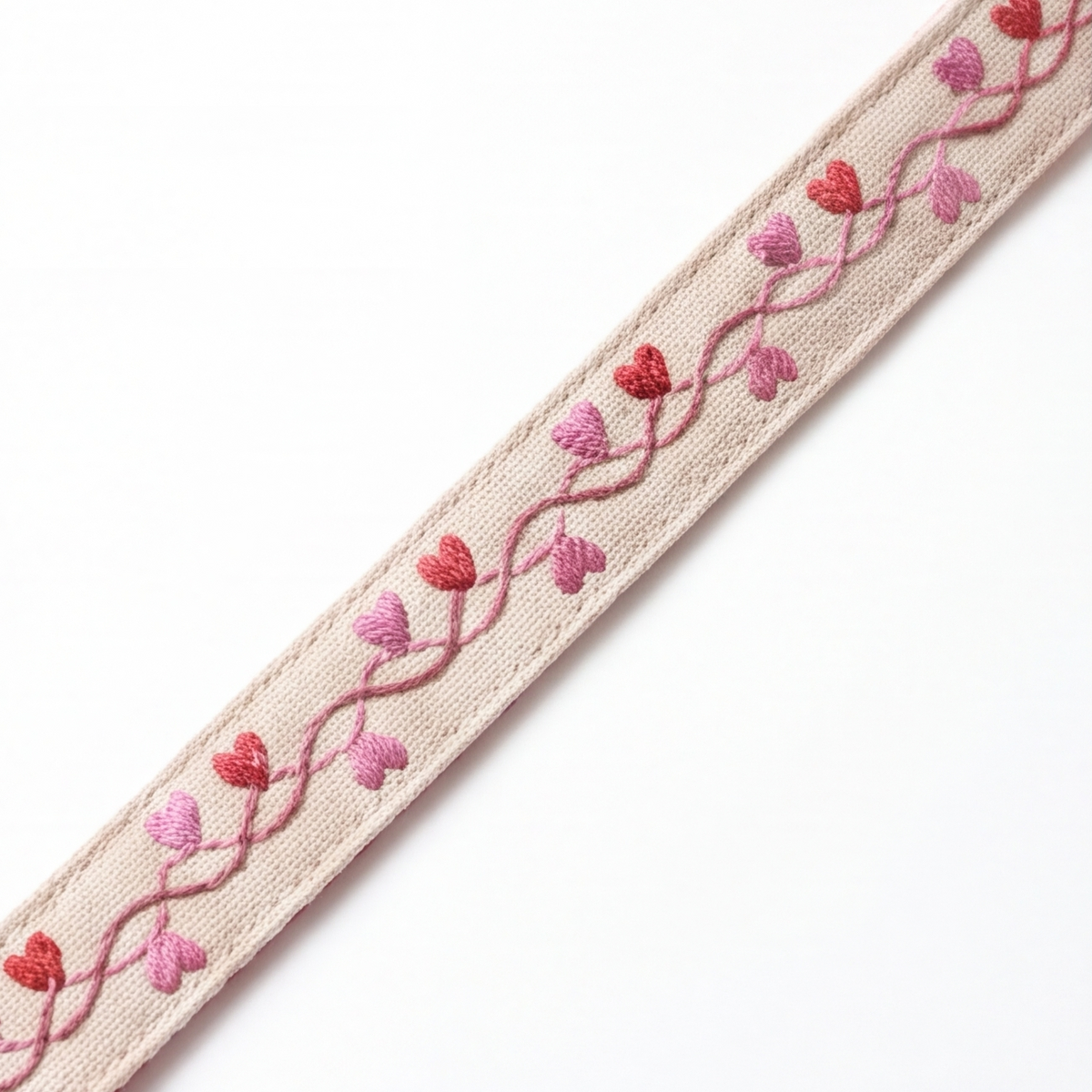 A close-up of a beige woven strap or trim featuring a wavy pink embroidered vine pattern with small red and pink heart motifs, shown diagonally on a plain white background.