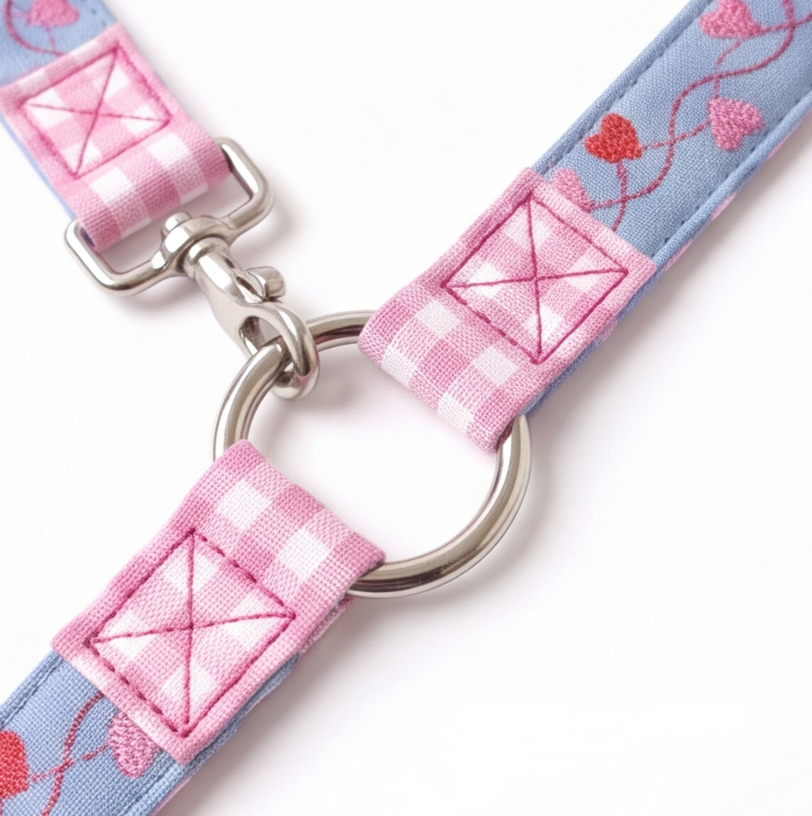 A close-up of a light blue fabric strap with pink gingham patches, embroidered pink hearts and swirls, and silver metal hardware including a ring and buckle, on a white background.