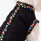A close-up of a black dog coat with colorful floral embroidery along the edges, worn by a small white dog against a plain white background.