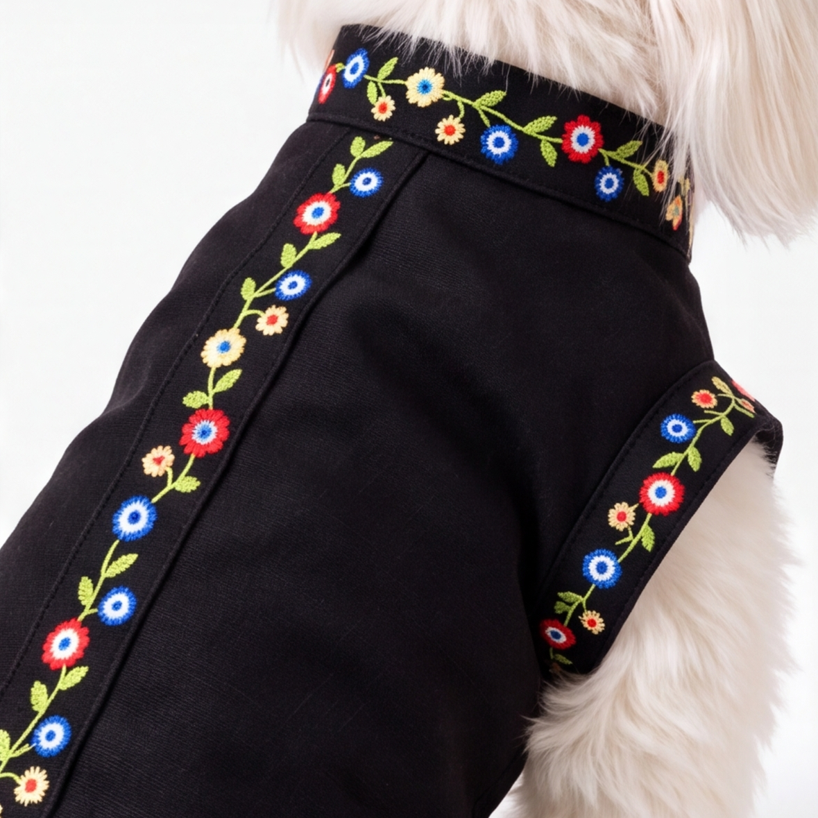 A close-up of a black dog coat with colorful floral embroidery along the edges, worn by a small white dog against a plain white background.