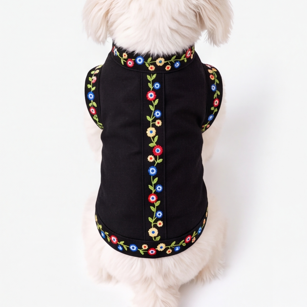 A small white dog viewed from behind wearing a black pet vest with colorful floral embroidery around the neckline, back, and hem, against a plain white background.