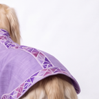 A close-up of a dog from behind wearing a lavender purple dog blanket with a geometric patterned border, set against a plain white background.