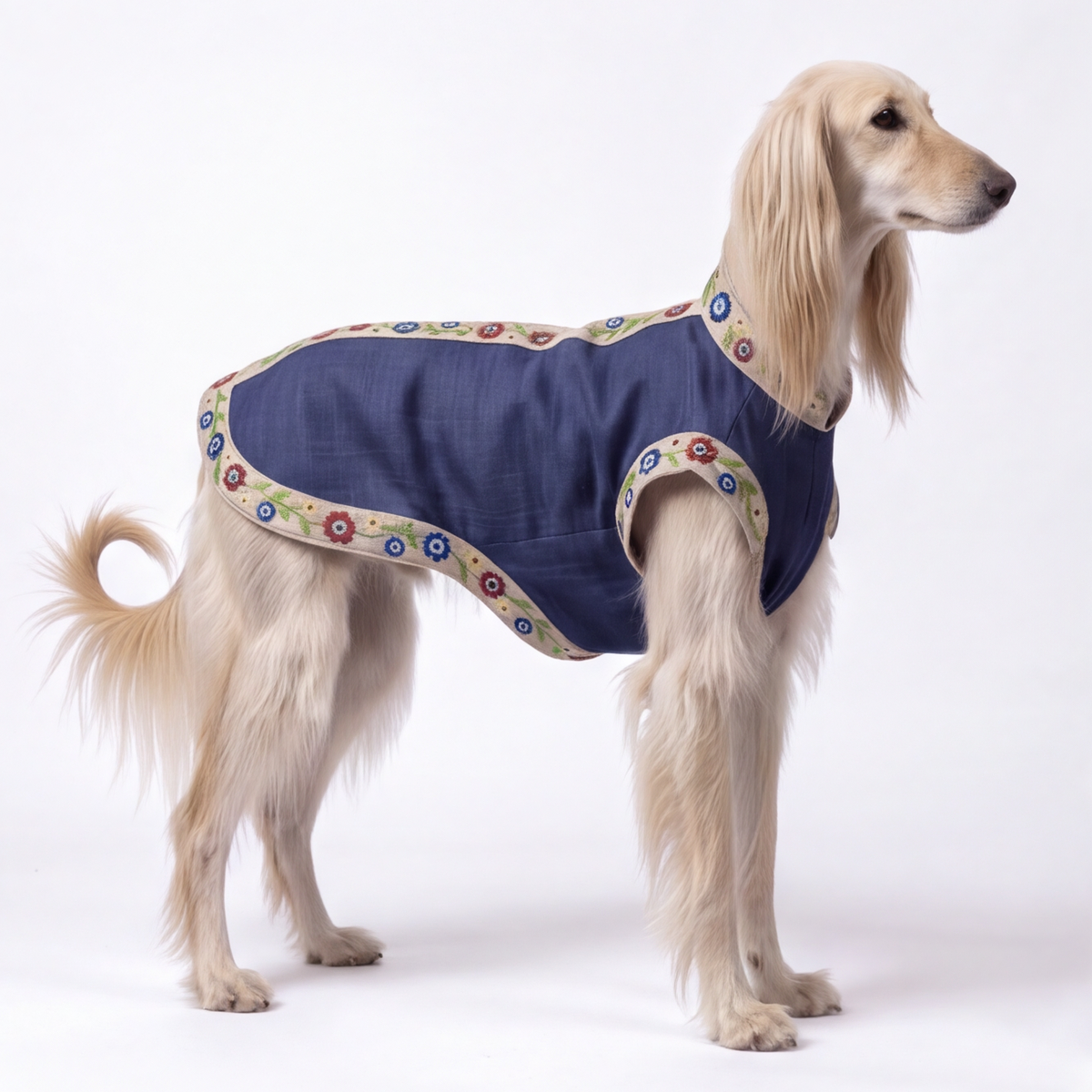 A long-haired light tan dog wearing a navy blue quilted pet jacket with colorful floral patterned trim, photographed against a seamless white studio background.