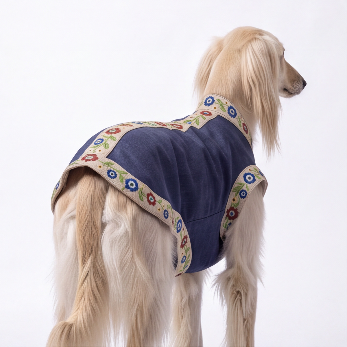 A long-haired light tan dog shown from the side and back wearing a navy blue dog coat with beige floral-patterned trim, photographed against a plain white studio background.